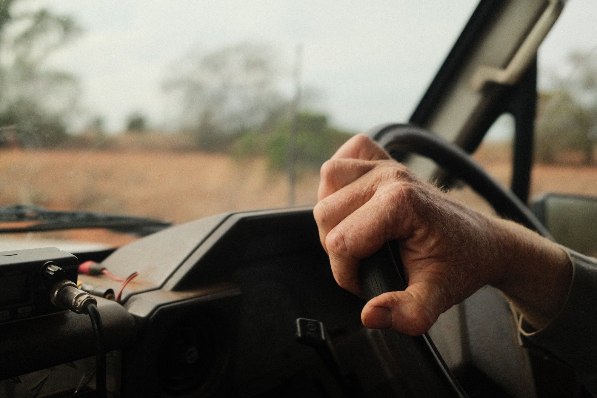 A weathered hand on the steering wheel of a work vehicle, with dry red-brown country and trees blurred through the windscreen.
